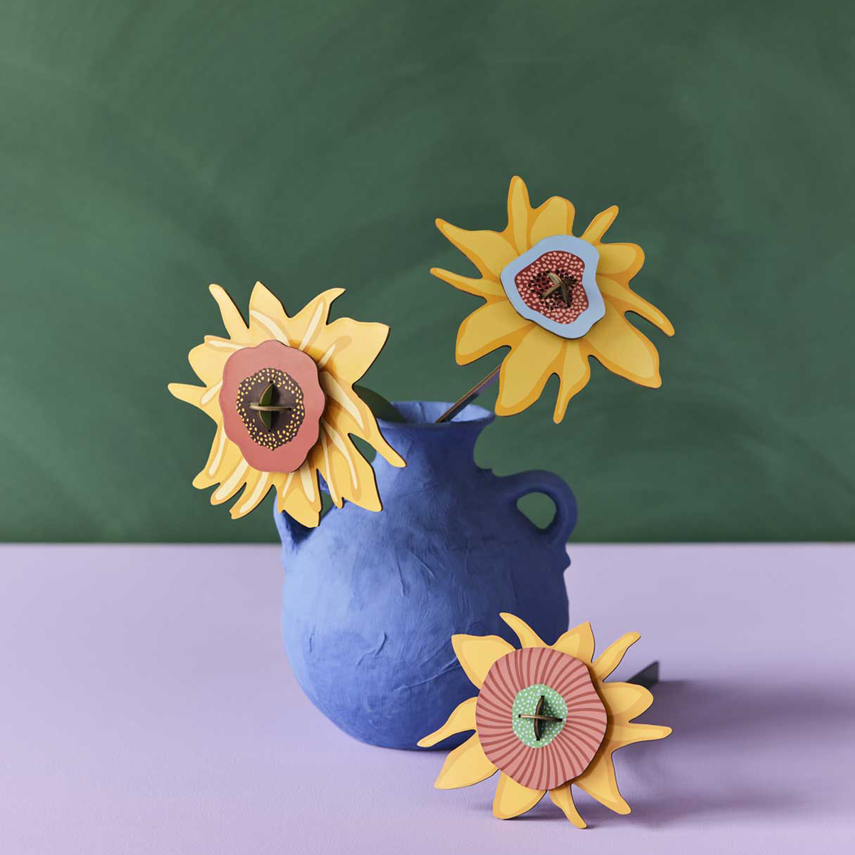 Yellow cardboard Van Gogh sunflowers in a blue vase against a green wall on a lilac table