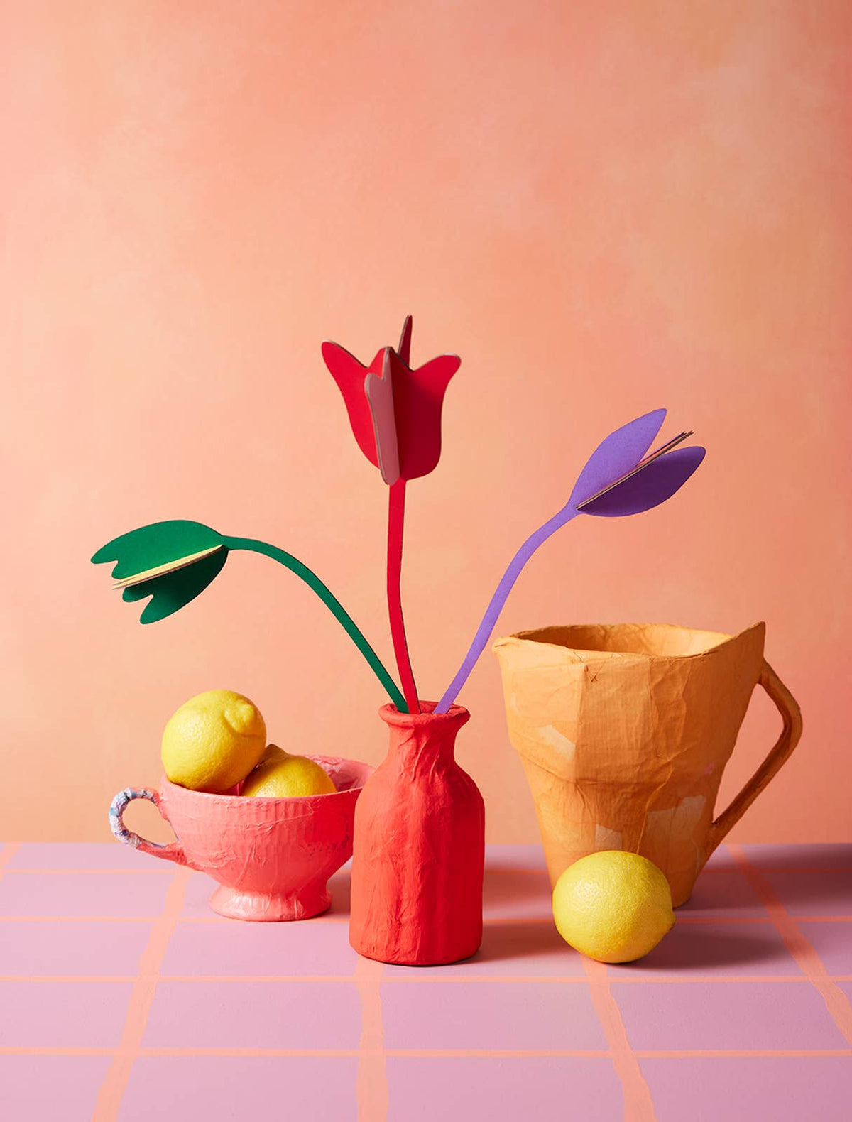 Red, purple and green cardboard tulips in a coral vase with lemons and a paper pitcher on a peach backdrop