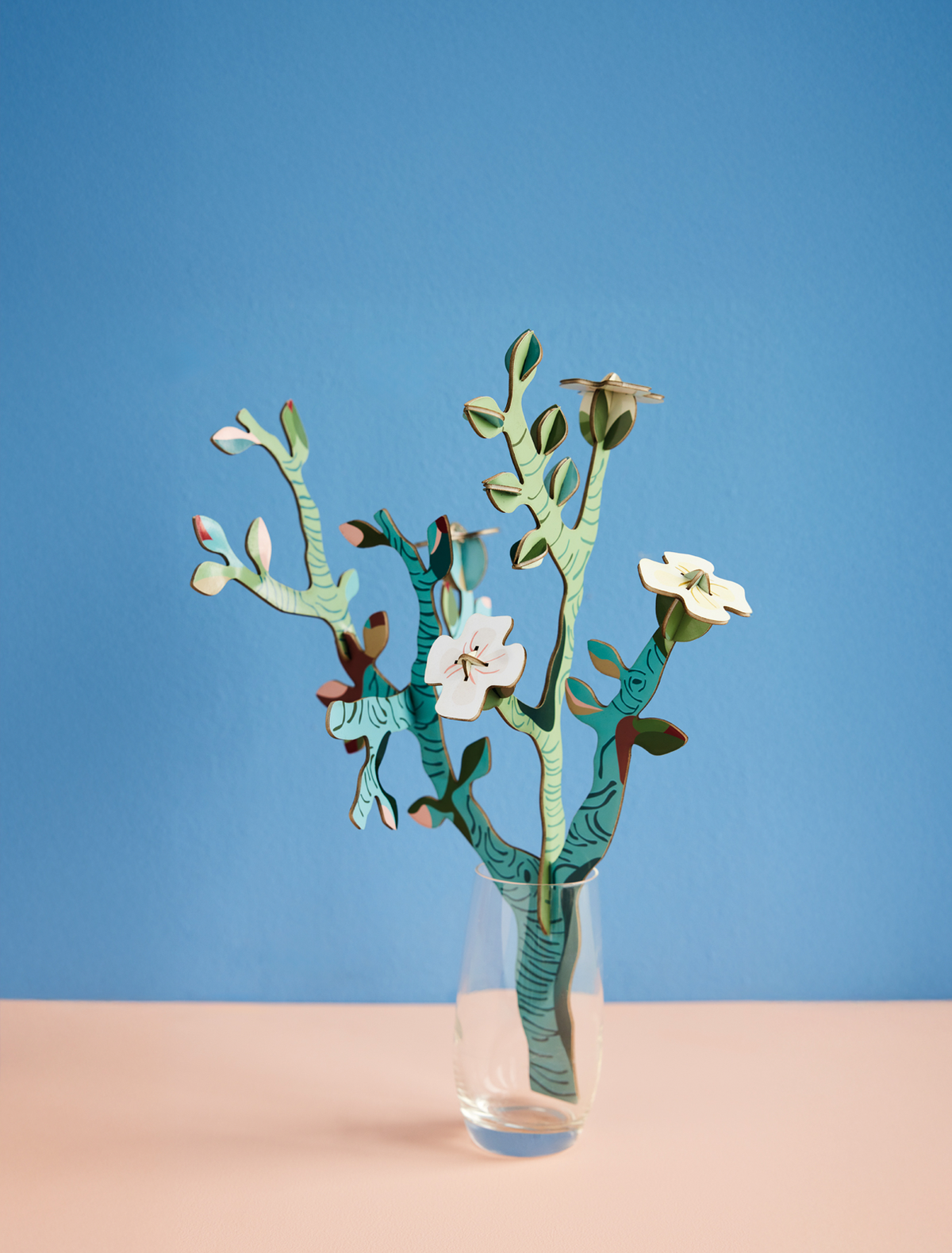 Cardboard almond blossom branches with white flowers in a clear glass vase against a blue wall